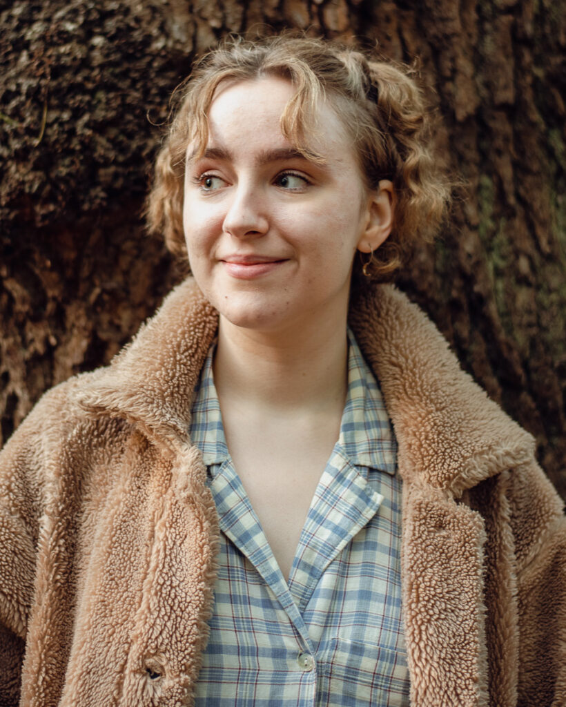 Zoe, a white person with short light brown hair. They are standing in front of a tree, smiling and looking to the left.