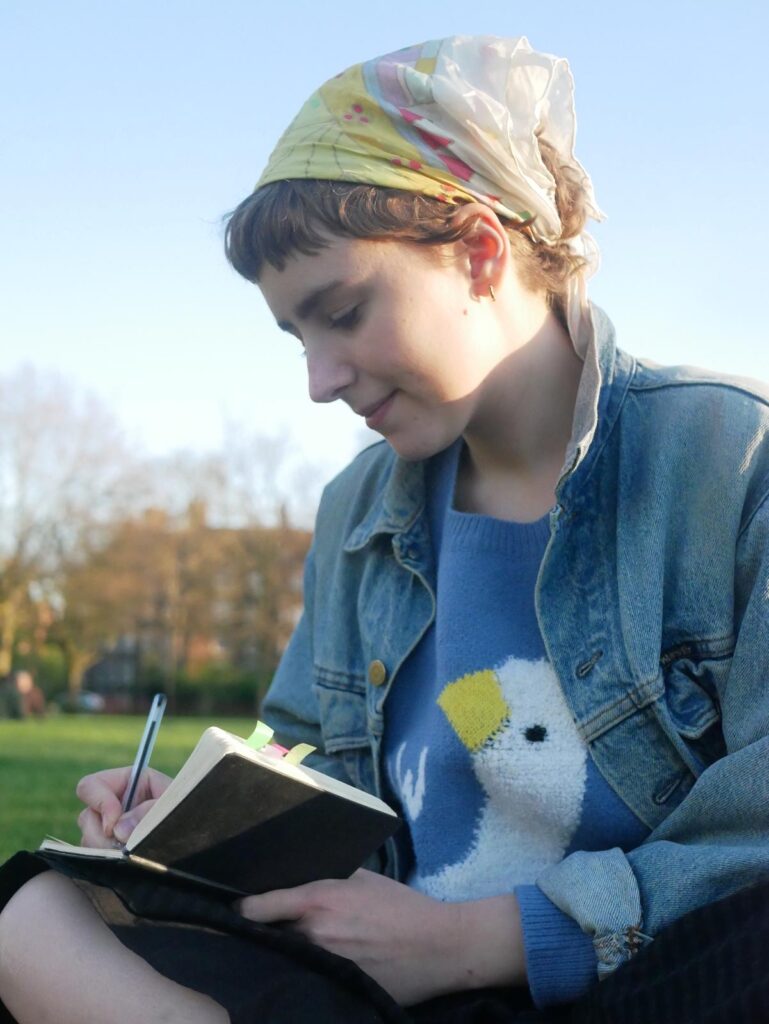 Zoe writes in a notebook, sitting in a park in the sun.